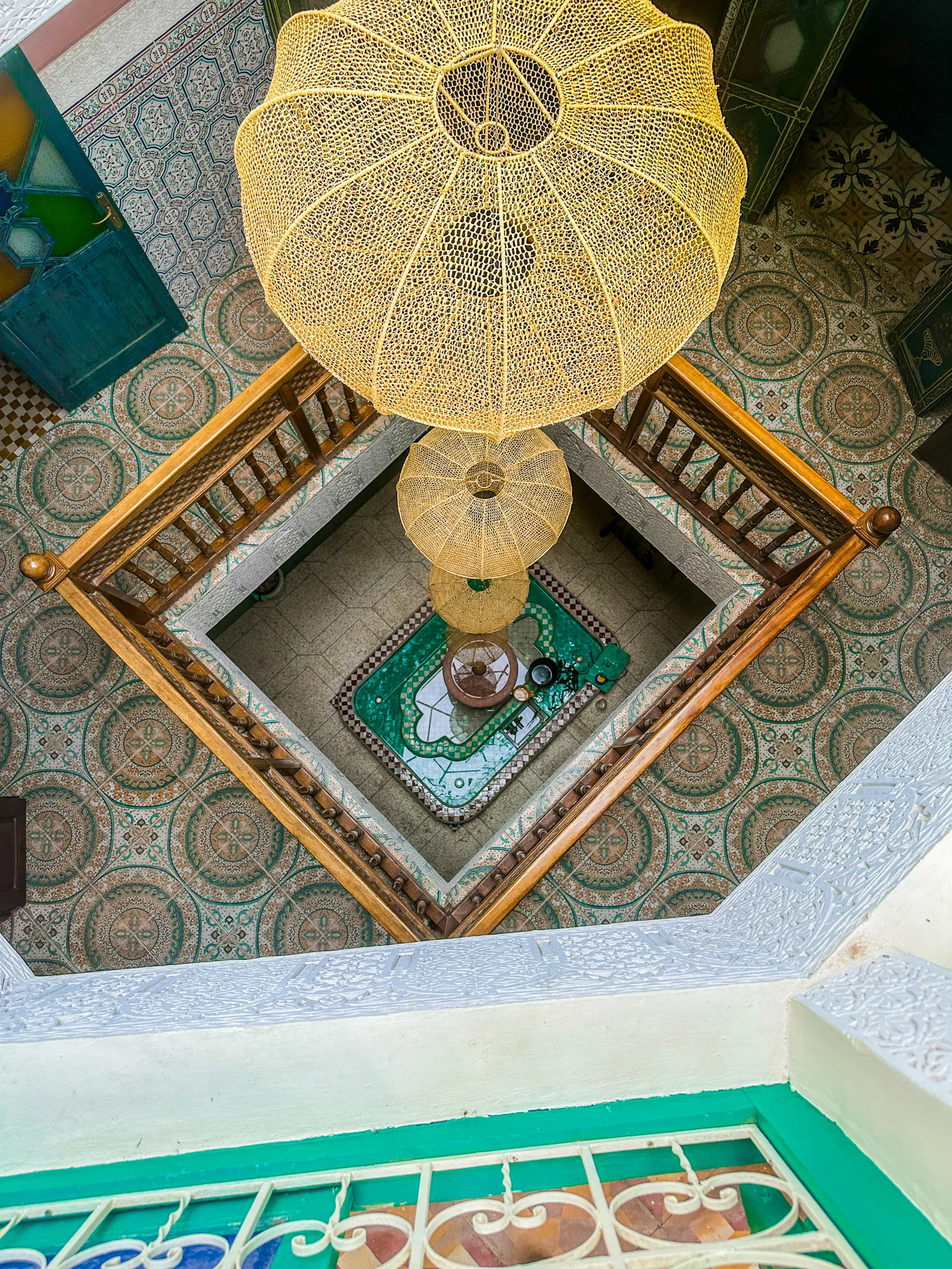 View from the first-floor gallery of Riad Tasneem down to the patio fountain, past woven pendant lamps and green zellige floors
