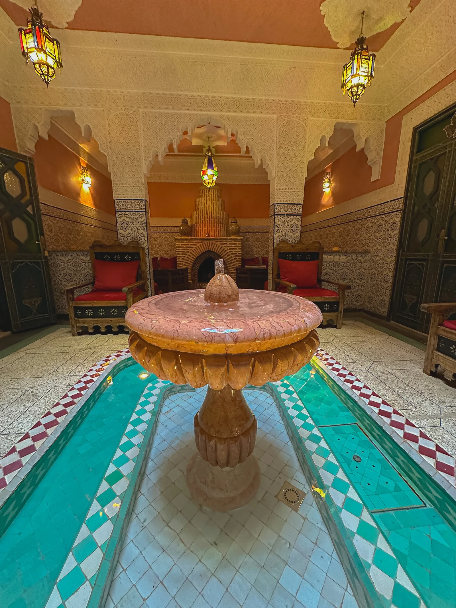 The patio of Riad Tasneem: pink marble fountain over a turquoise zellige basin, carved stucco arches, stained-glass lanterns and a traditional fireplace