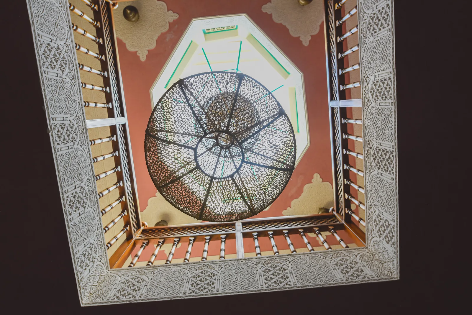 Looking up through the lightwell of Riad Tasneem: carved stucco frames, wooden balustrades and a woven lantern under the octagonal skylight