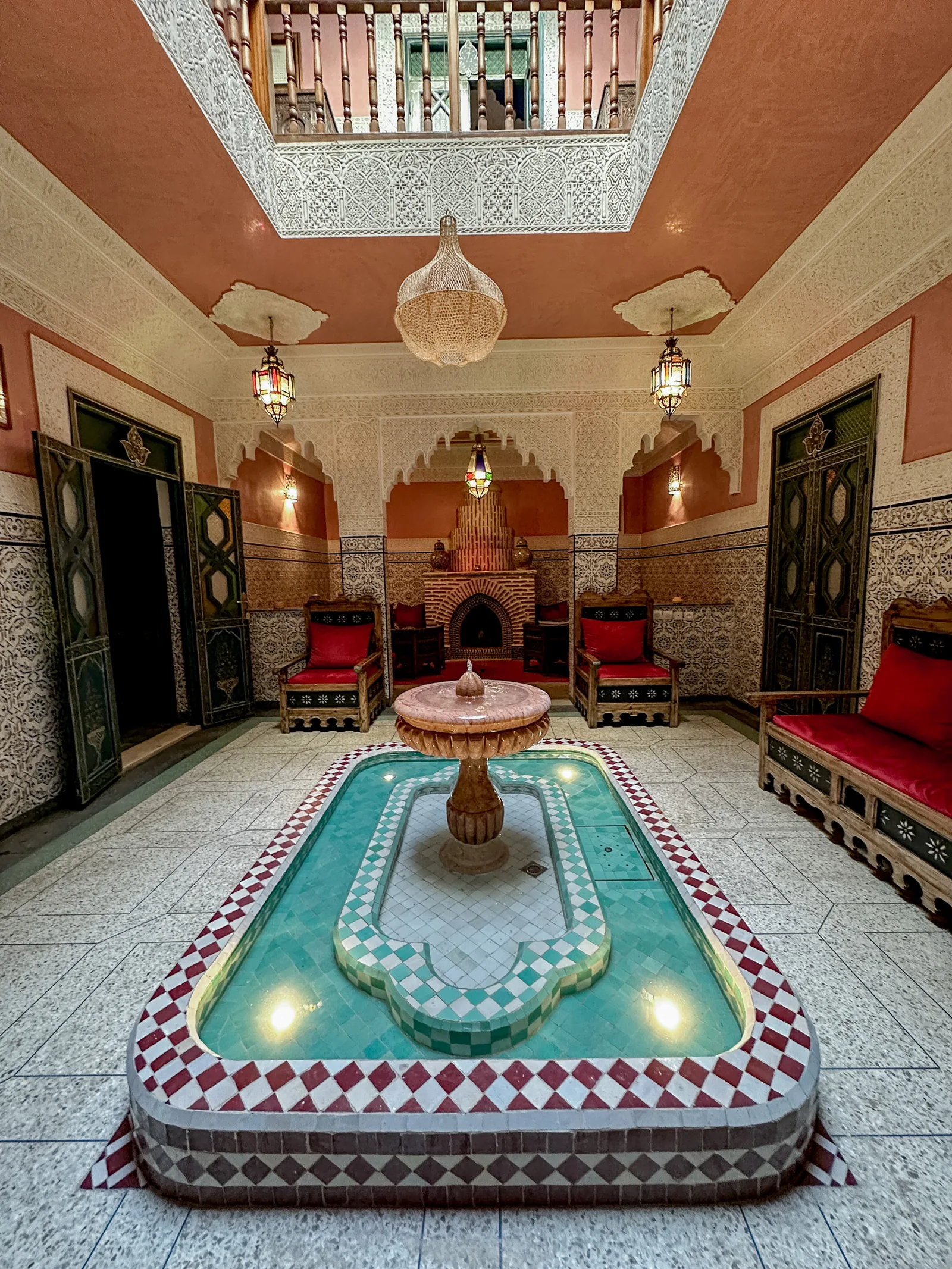 The patio of Riad Tasneem: marble fountain over a turquoise zellige basin, carved stucco arches, traditional fireplace and red-cushioned seats under the gallery