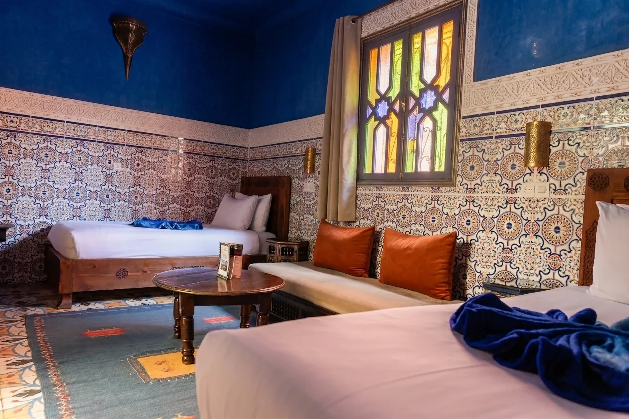 Safaa room from a side angle: foreground bed with blue throw, carved wooden frame, view onto stained-glass window and second bed beyond