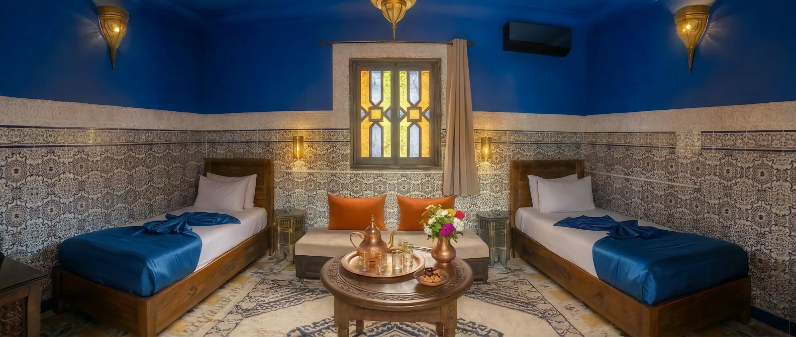 Panoramic view of Safaa room at Riad Tasneem: two single beds left and right, central seating bench under a stained-glass window, deep blue tadelakt ceiling, zellige tiled walls and Berber rug