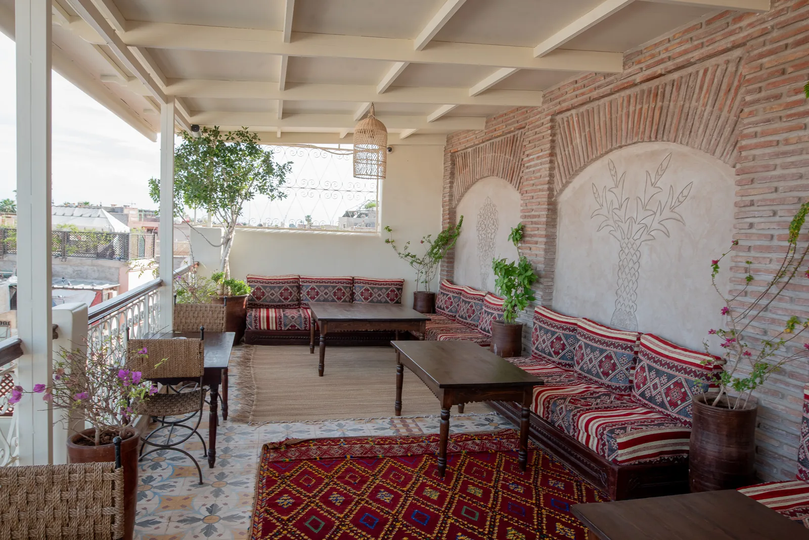 Covered rooftop lounge at Riad Tasneem: kilim sofas along palm reliefs in the brick wall, Berber rugs and views over the rooftops of the medina